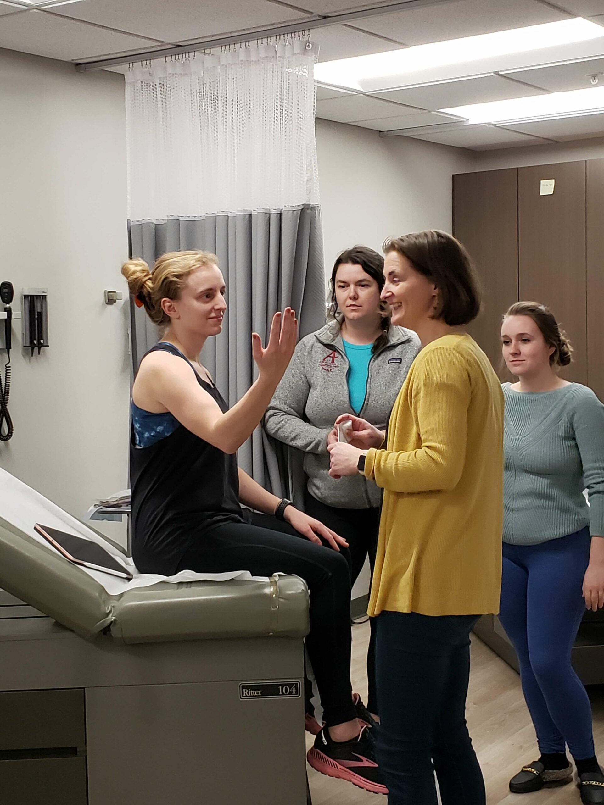 Professor and 3 students around a clinical bay learning a lesson. One student is sitting on the bed and holding up her hand